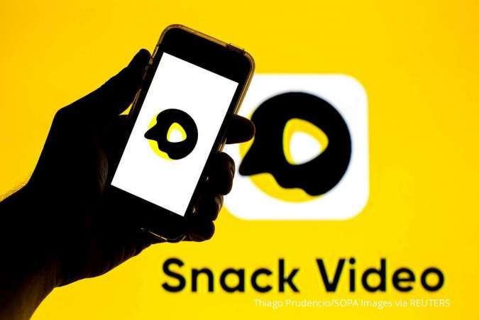 Sebuah tangan yang memegang smartphone menampilkan logo Snack Video, dengan logo yang sama dan teks "Snack Video" ditampilkan di latar belakang layar kuning.