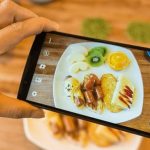 Seseorang memotret sepiring sarapan dengan ponsel pintar. Sepiring itu berisi kiwi, irisan jeruk, pisang, telur dadar, dan sosis.