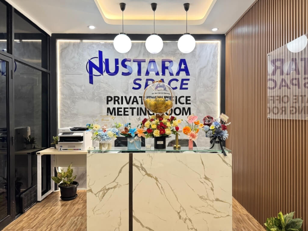 Area resepsionis di Nustara Space dengan meja marmer, rangkaian bunga warna-warni, dan papan bertuliskan "Private Office Meeting Room" di dinding belakang.